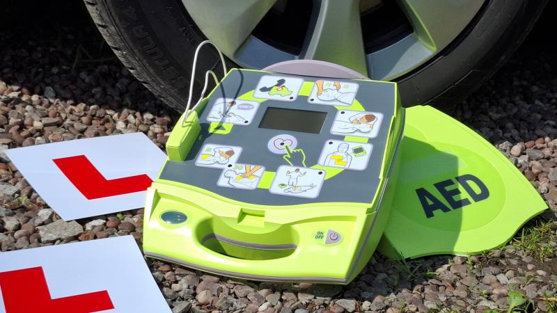 Automated external defibrillator placed on the ground next to a car tyre, with L‑plates nearby.